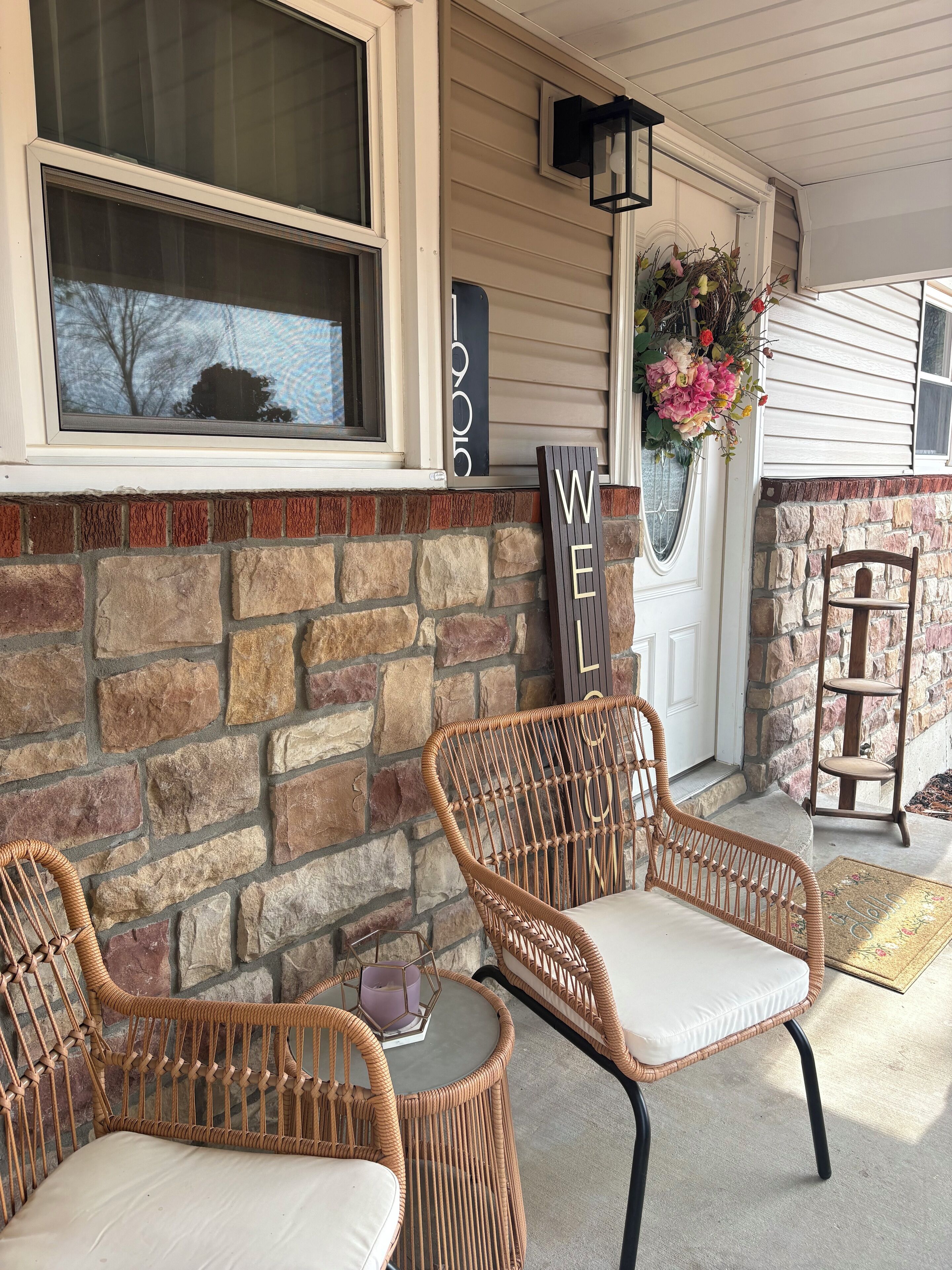 A porch with two wicker chairs, a small table, a welcome sign, and a flower arrangement.