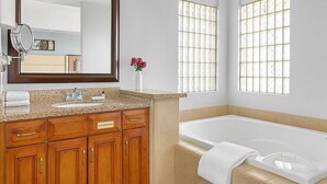 Un baño con encimera de granito, armarios de madera, lavamanos blanco, un espejo grande, una bañera blanca y una ventana con vistas al exterior.
