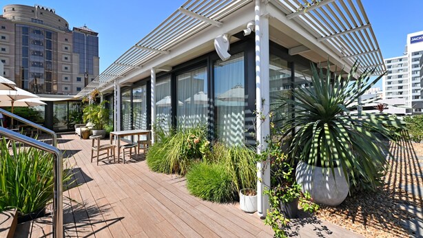 Uma área de estar moderna ao ar livre com um deck de madeira, plantas em vasos e um edifício com paredes de vidro.