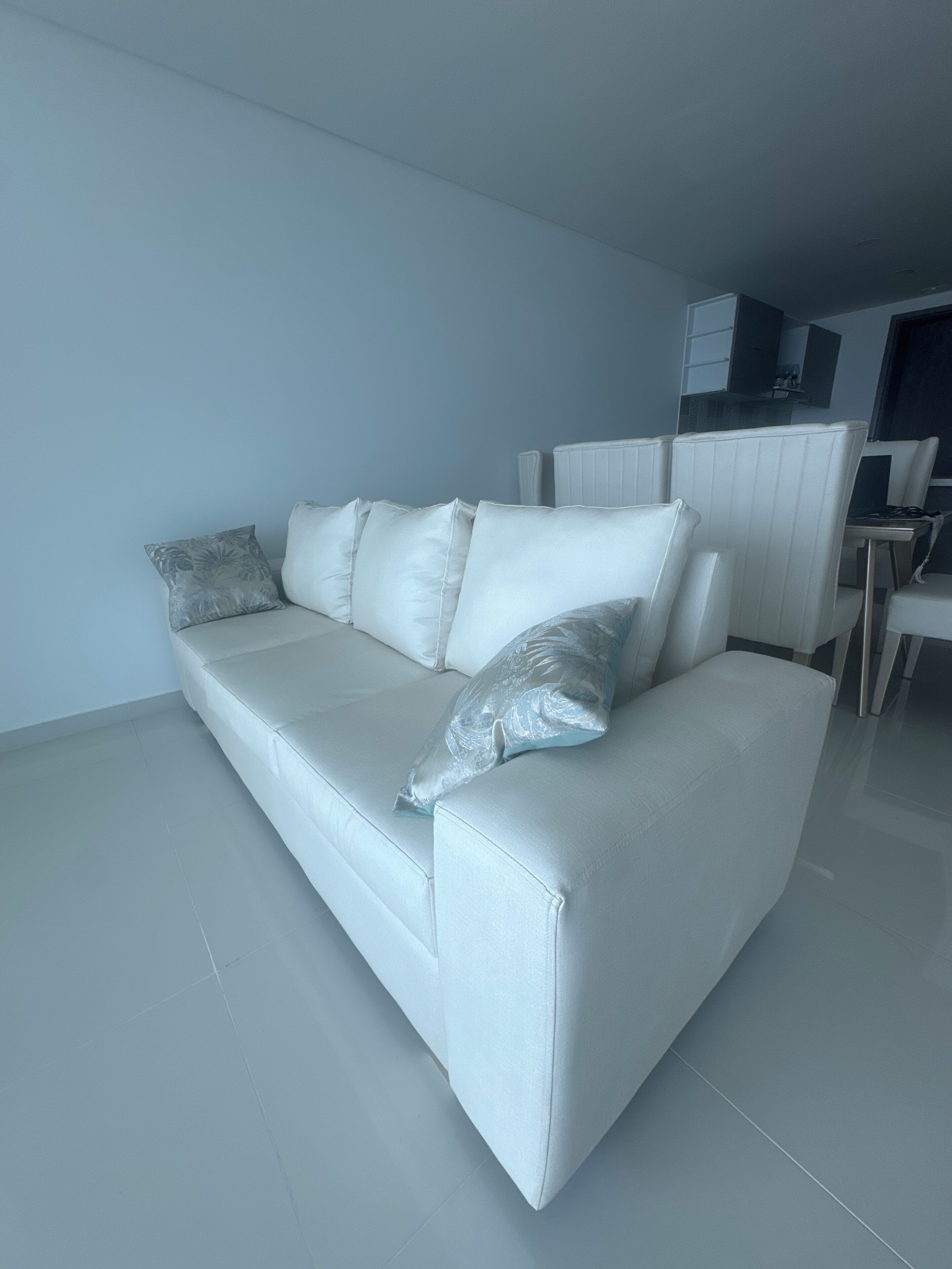 A modern living room with a white sofa, a dining area with a table and chairs, and a kitchen area with cabinets and a countertop.