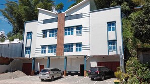 Un edificio de varios pisos con una chimenea de ladrillo en el centro, múltiples ventanas y un estacionamiento con dos coches.