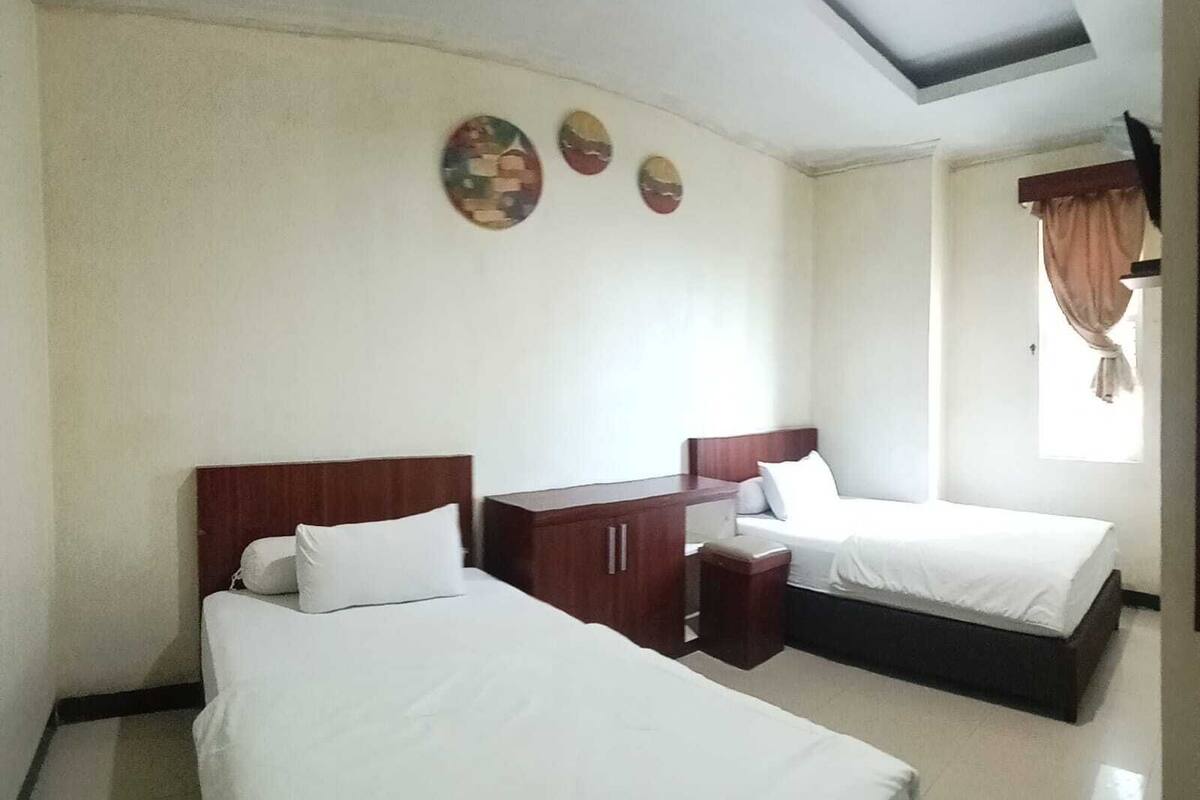 A hotel room with two beds, a wooden nightstand, a small table, and a window with curtains.