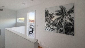 Un intérieur moderne avec un mur blanc, un grand tableau représentant un palmier en noir et blanc, et une vue sur un balcon extérieur, accessible par une porte.