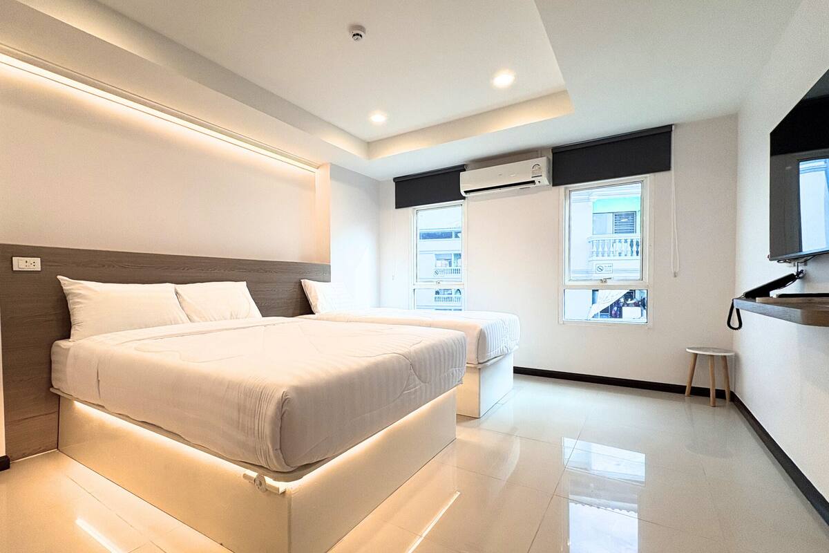 A modern hotel room with a large bed, a small bedside table, a built-in air conditioning unit, and a window with a view of a building.
