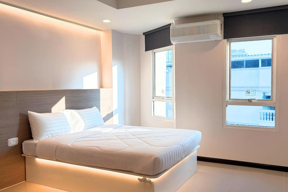 A modern hotel room with a bed, a window with a view, an air conditioning unit, and a wall-mounted outlet.