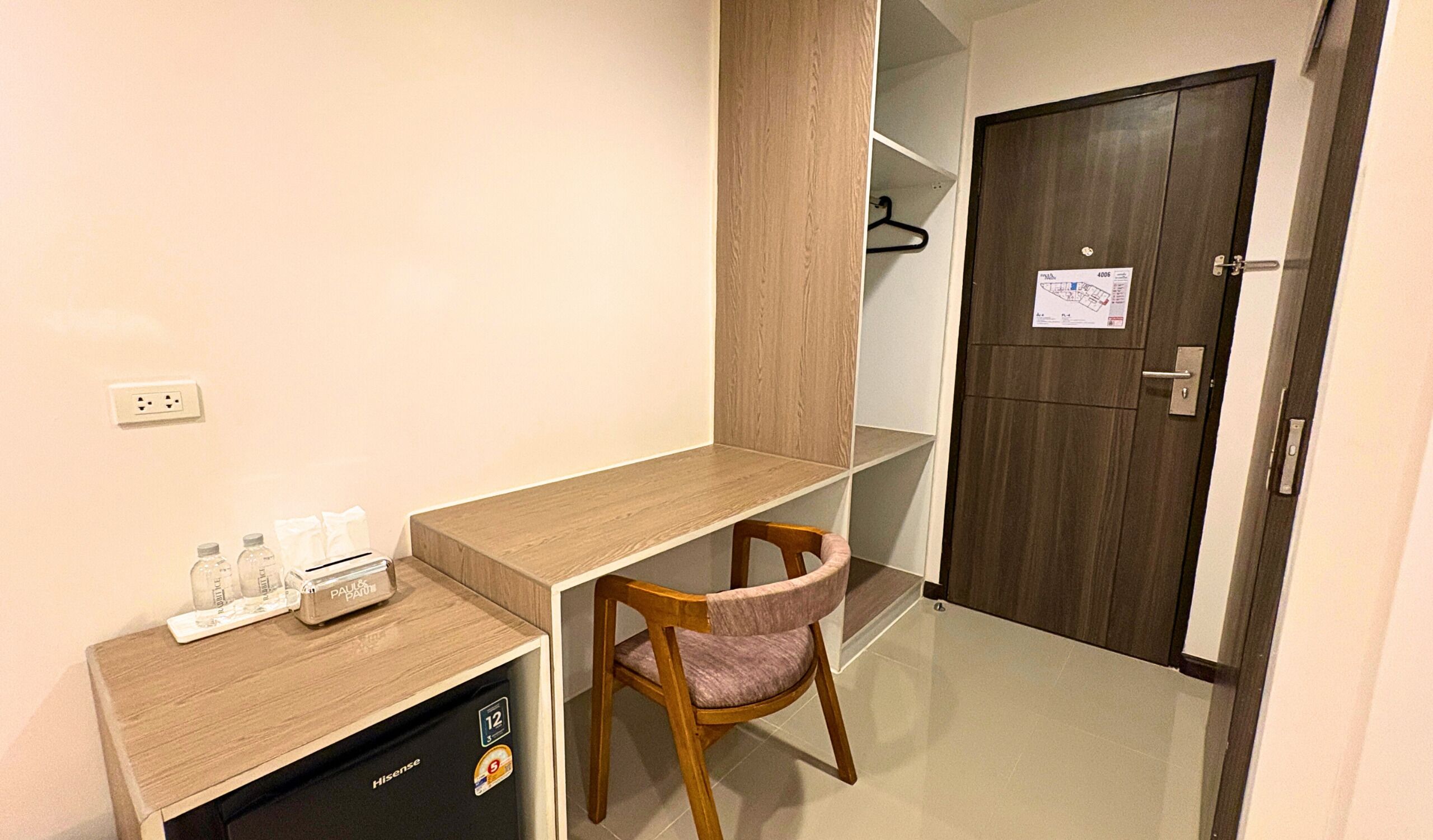 Habitación pequeña con escritorio de madera, una silla, un armario bajo y una toma de corriente, además de una puerta con un aviso.