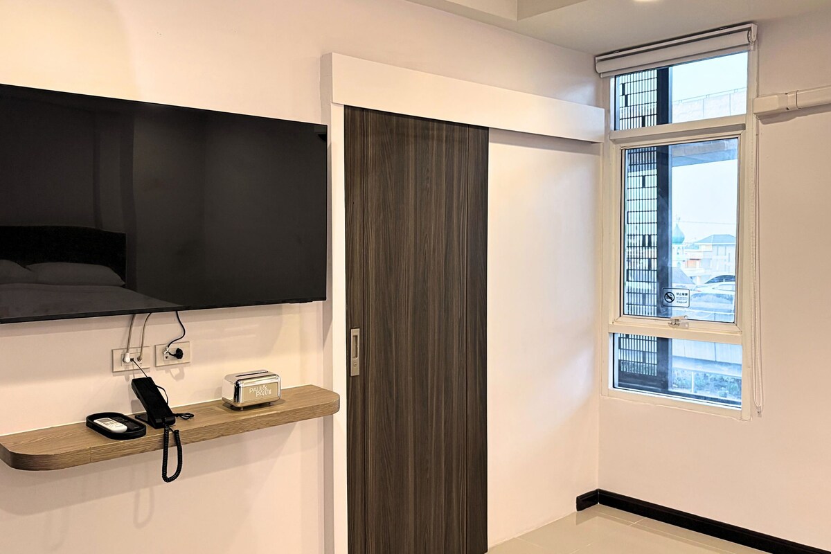 A hotel room with a flat-screen TV mounted on the wall, a wooden shelf with a telephone and a toaster, and a window with a view of buildings.