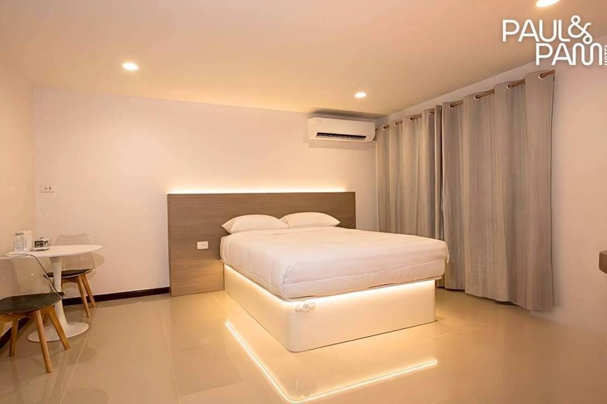 A modern hotel room with a bed, bedside table, chair, and an air conditioning unit.