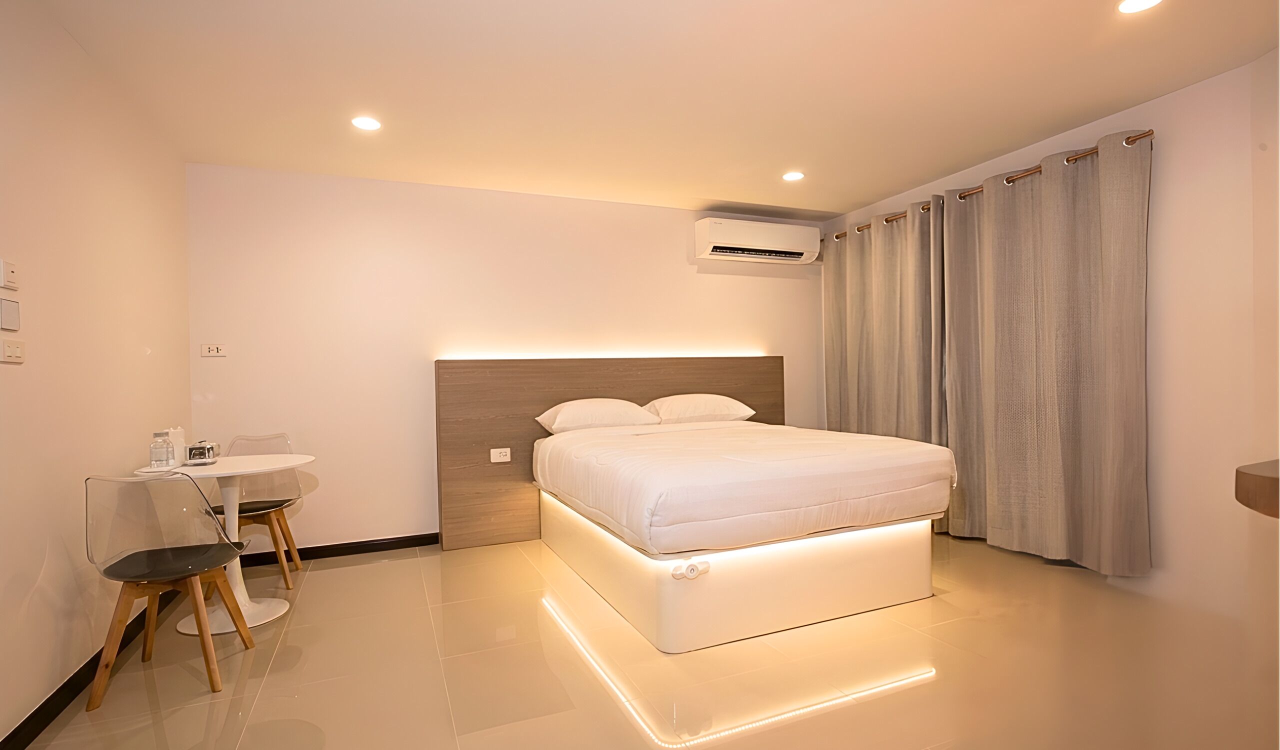 A modern hotel room with a bed, bedside table, chair, and an air conditioning unit.