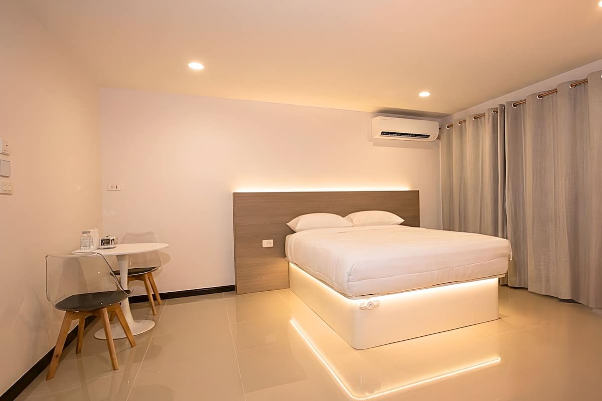 A modern hotel room with a bed, bedside table, chair, and an air conditioning unit.