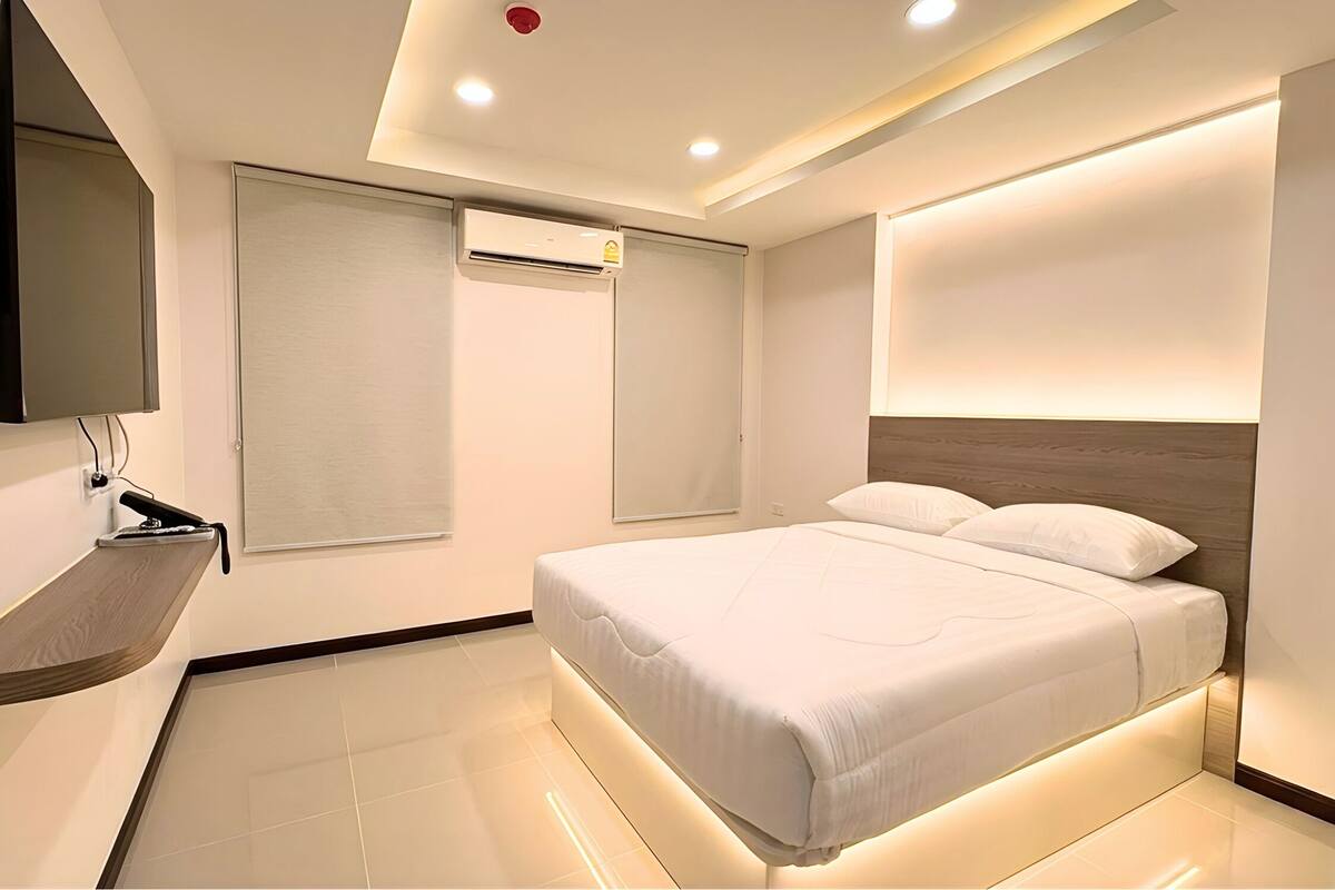 A modern hotel room with a bed, a flat-screen TV, a wall-mounted air conditioner, and a ceiling light.