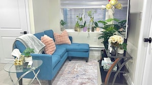 Ein Wohnzimmer mit blauem Sofa, einem Glastisch, einem kleinen Beistelltisch, einem gemusterten Teppich und verschiedenen topfpflanzen.