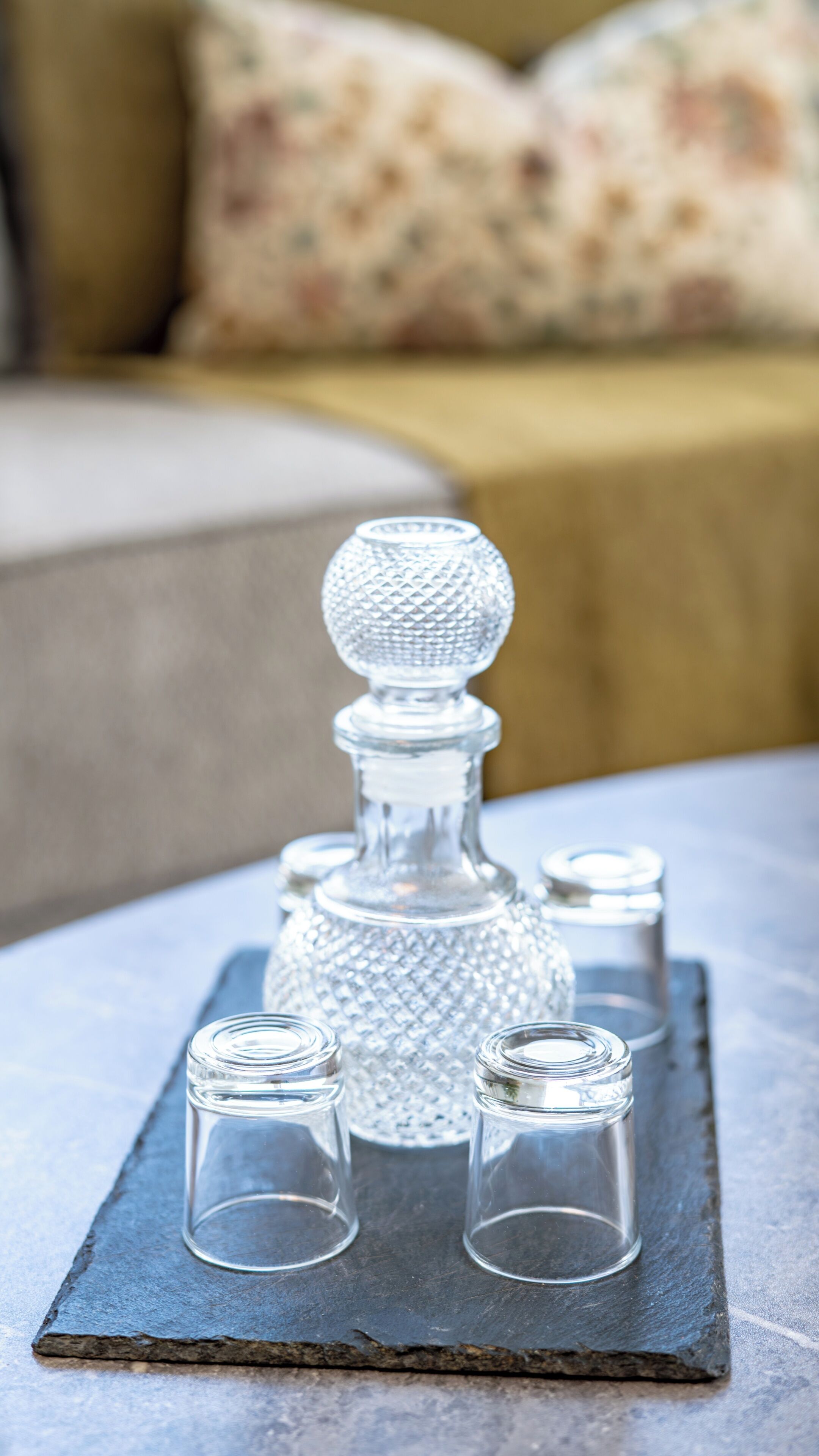 A clear glass decanter with a textured stopper and four small glasses on a slate tray.