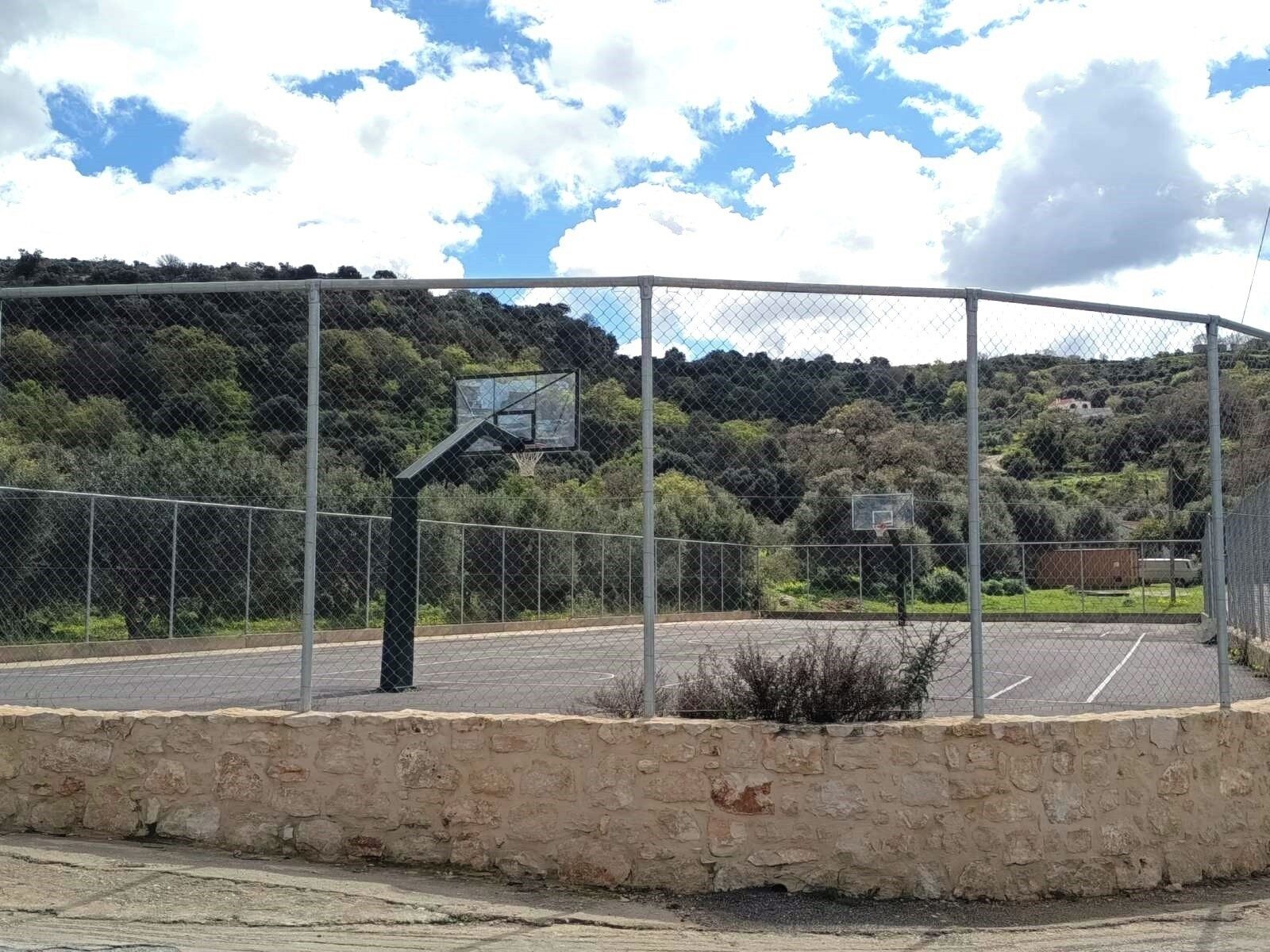 Un terrain de basketball en plein air, délimité par une clôture en treillis métallique et doté d’une base en muret de pierres, entouré d’arbres et de collines.