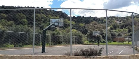 An outdoor basketball court enclosed by a chain-link fence with a stone wall base, surrounded by trees and hills.