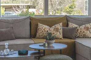 A living room with a sofa, a round coffee table, and a window with a view of trees and a building. - Thyme Aroma - Quiet Nature Patio (Gonia, Rethymno Crete)