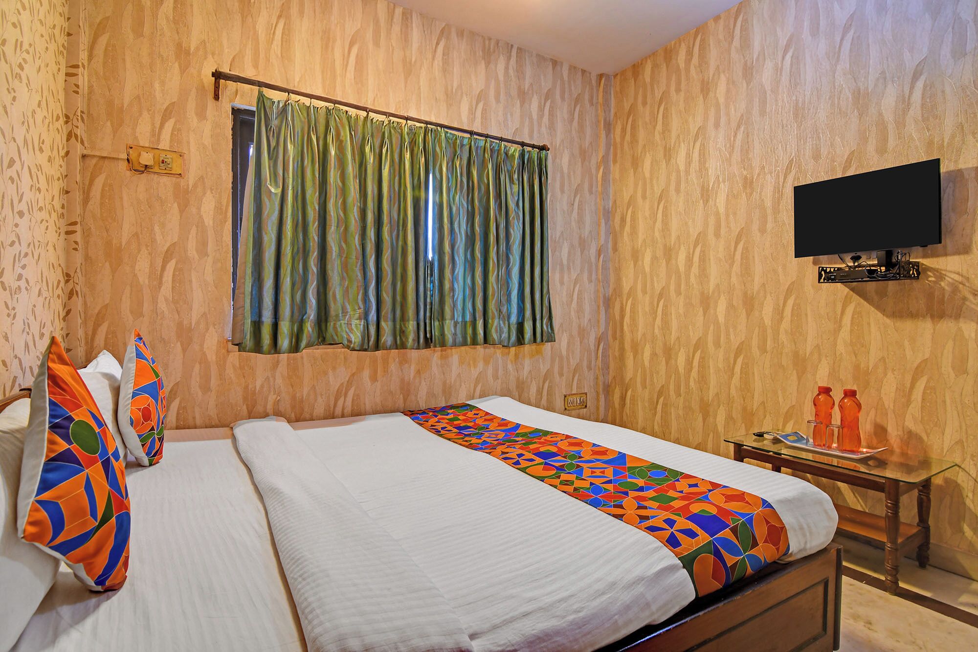 A hotel room with a bed, a television, a small table with bottles, and a window with curtains.