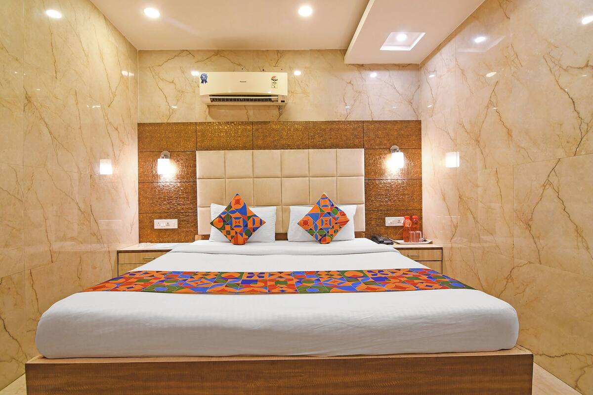 A hotel room with a marble wall, a bed with a colorful geometric bedspread, a headboard with built-in lighting, and an air conditioning unit.