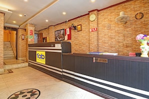 A hotel reception area with a reception desk, a clock, a wall-mounted fan, and a staircase leading to an upper floor.