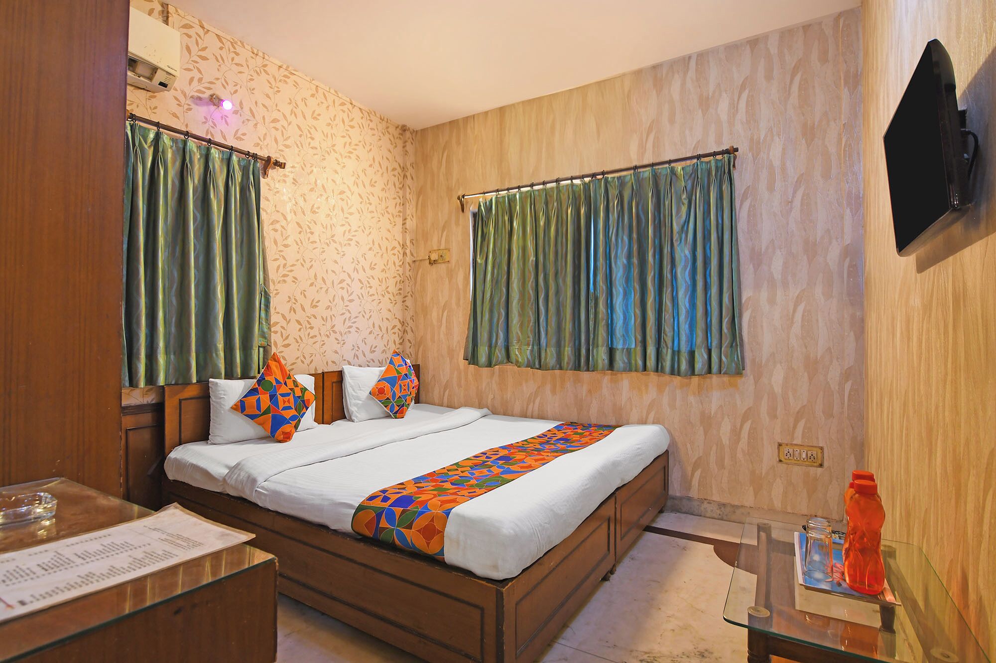 Een hotelkamer met een houten bed, twee kussens, een glazen tafel en een aan de muur bevestigde televisie.