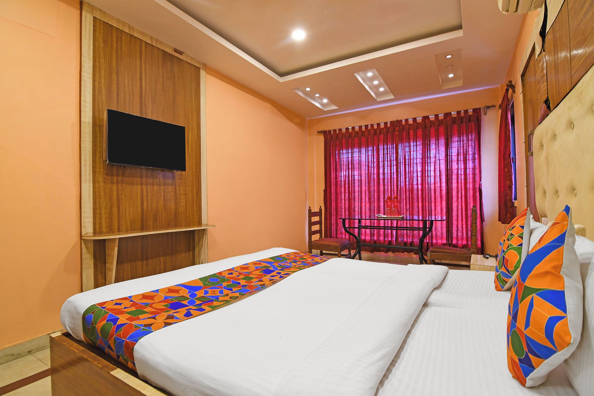 A hotel room with a bed, a TV mounted on the wall, a desk with a chair, and a window with curtains.