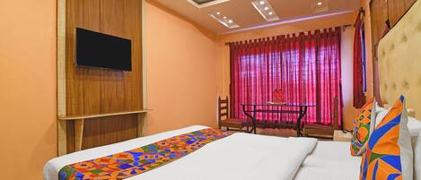 A hotel room with a bed, a TV mounted on the wall, a desk with a chair, and a window with curtains.
