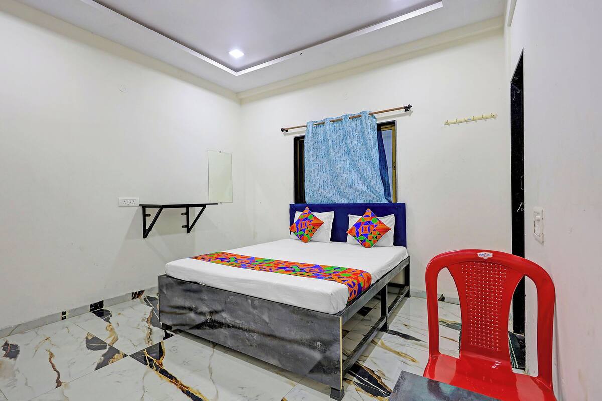 A small, clean room with a bed, a red chair, a window with a blue curtain, and a wall-mounted mirror.