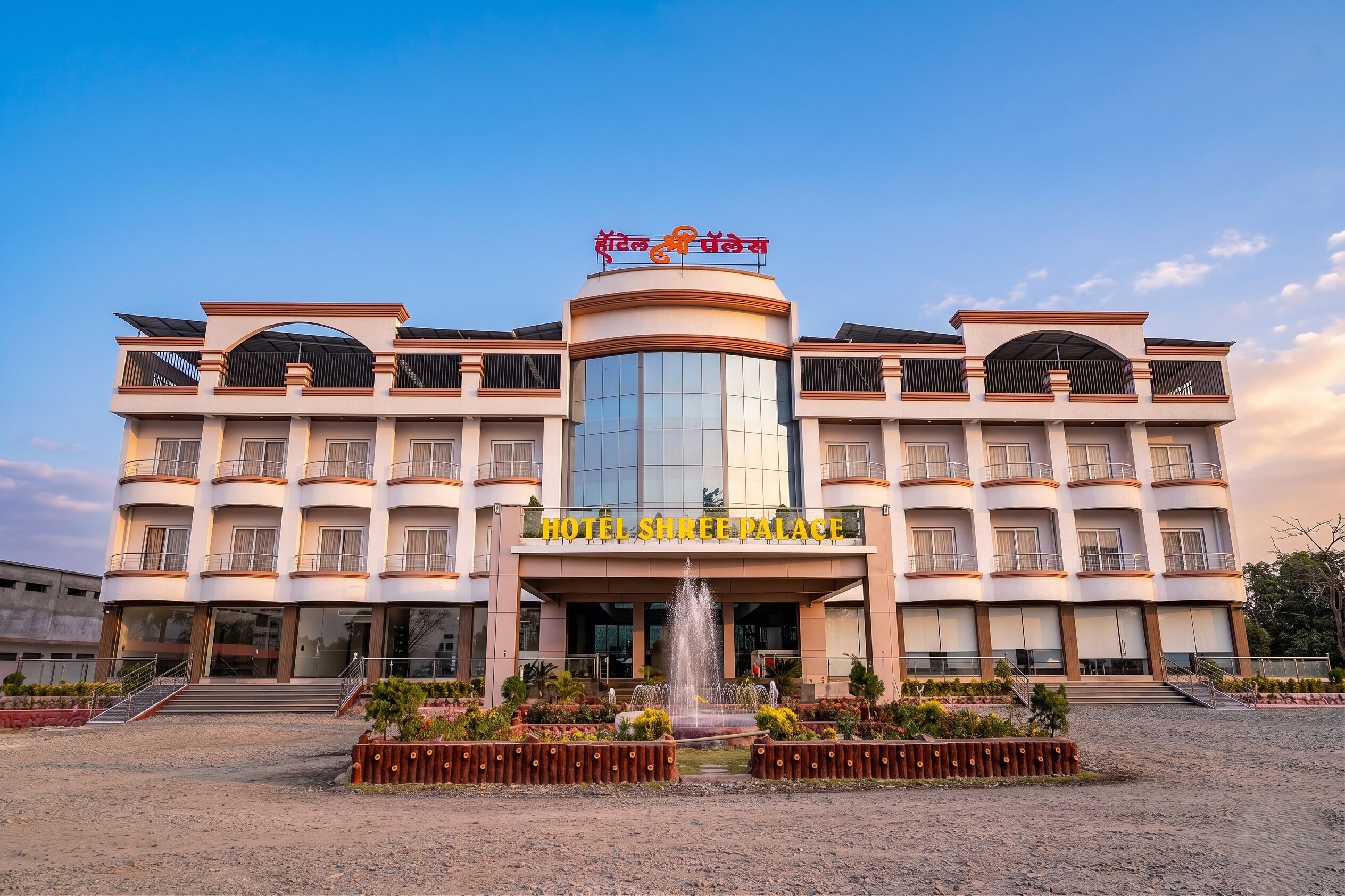 Un hotel de varios pisos con una fuente central, grandes ventanales y un cartel que dice 'HOTEL SHREE PALACE'.