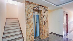 Um eixo de elevador revestido em mármore com porta de elevador azul, escada com degraus brancos e uma parede lisa.
