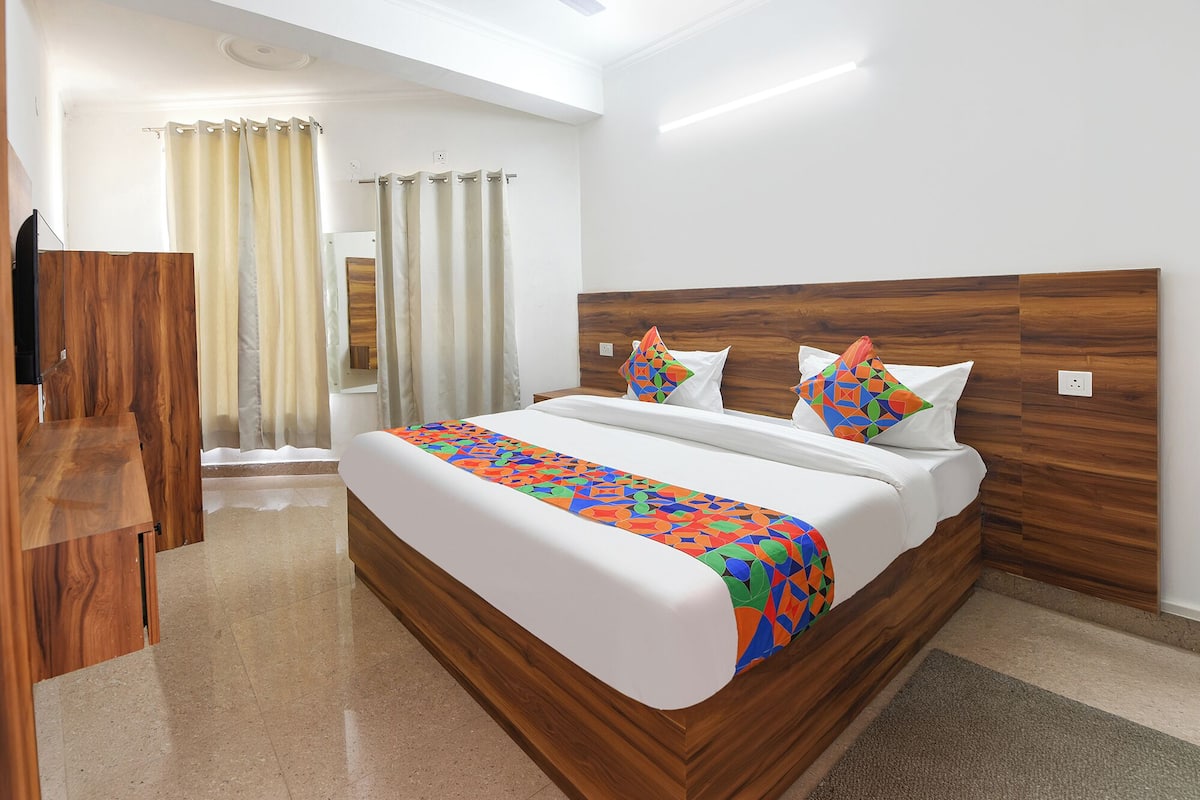 Ein Hotelzimmer mit Holzkopfteil, einem Bett mit weißer Bettwäsche, farbenfrohen geometrischen Kissen und Blick auf ein Badezimmer mit Dusche und Vorhängen.