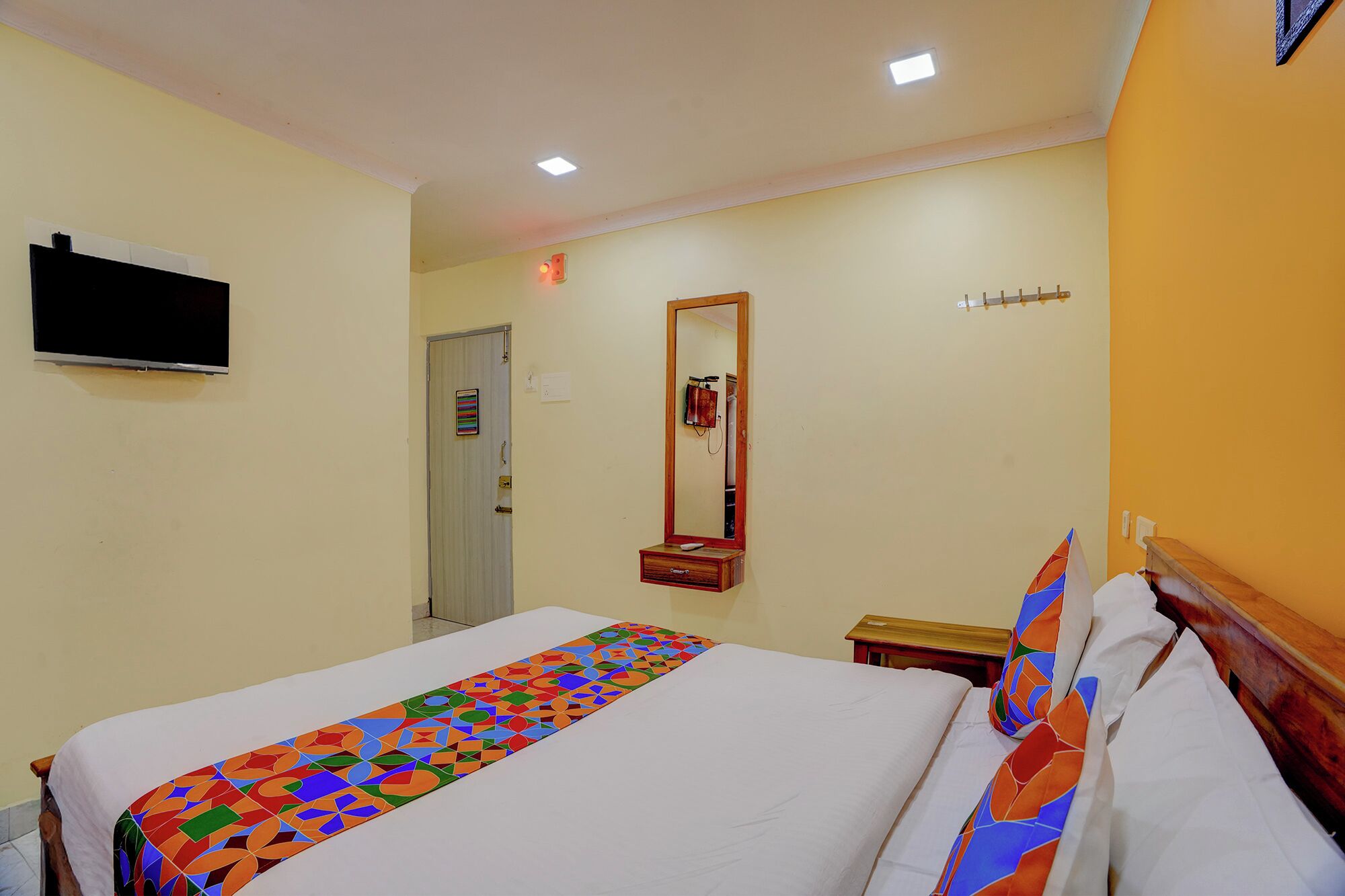 Habitación de hotel con una cama, un televisor montado en la pared, una puerta, un espejo y una mesita.