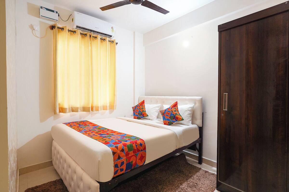 Un dormitorio con cama, ventilador de techo, unidad de aire acondicionado, puerta de madera y ventana con cortinas.