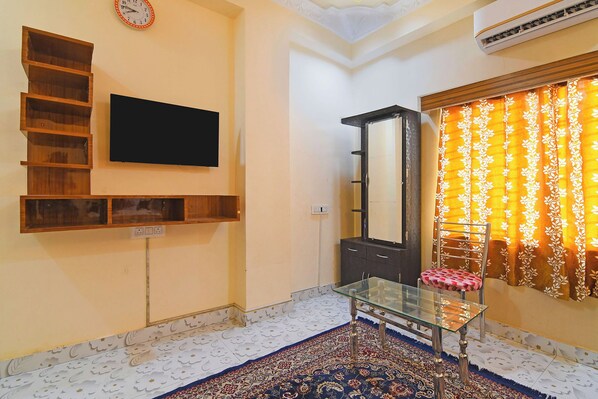 A living room with a TV, wooden shelves, a glass coffee table, a large mirror, a patterned rug, and a window with curtains. - Via Safehome Guest House (Barasat)
