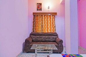 A living room with a brown sofa, a glass coffee table, patterned curtains, and a wall-mounted light fixture. - Via Safehome Guest House (Barasat)