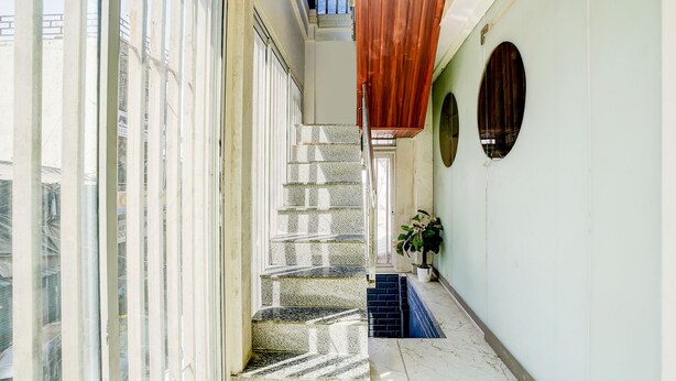 A narrow hallway with a staircase leading to an upper floor, large windows on one side, and a door with a wooden panel on the other. - Fabhotel Nm (Mumbai)