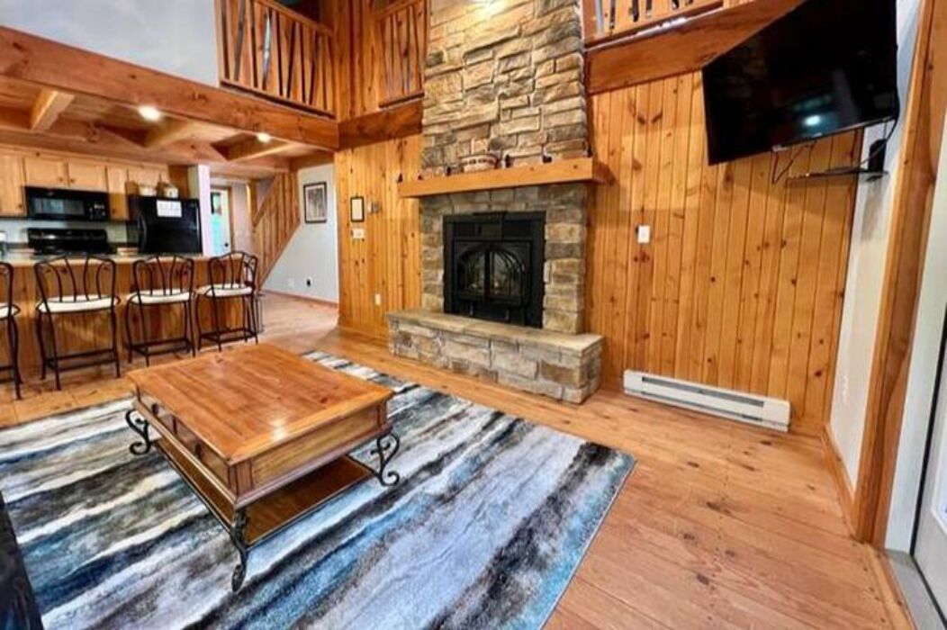 A wooden interior with a stone fireplace, a dining area with chairs, and a kitchen in the background.