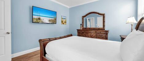 Un dormitorio con cama, ventilador de techo, cómoda de madera, espejo y televisor montado en la pared.