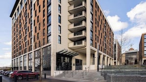 Un immeuble moderne à plusieurs étages, doté d’une entrée imposante, d’un espace abrité et d’un parking où sont stationnées des voitures.
