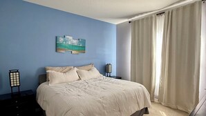 Ein Schlafzimmer mit einem Bett, einem Nachttisch, einer Lampe, einem Gemälde und einem Fenster mit Vorhängen.