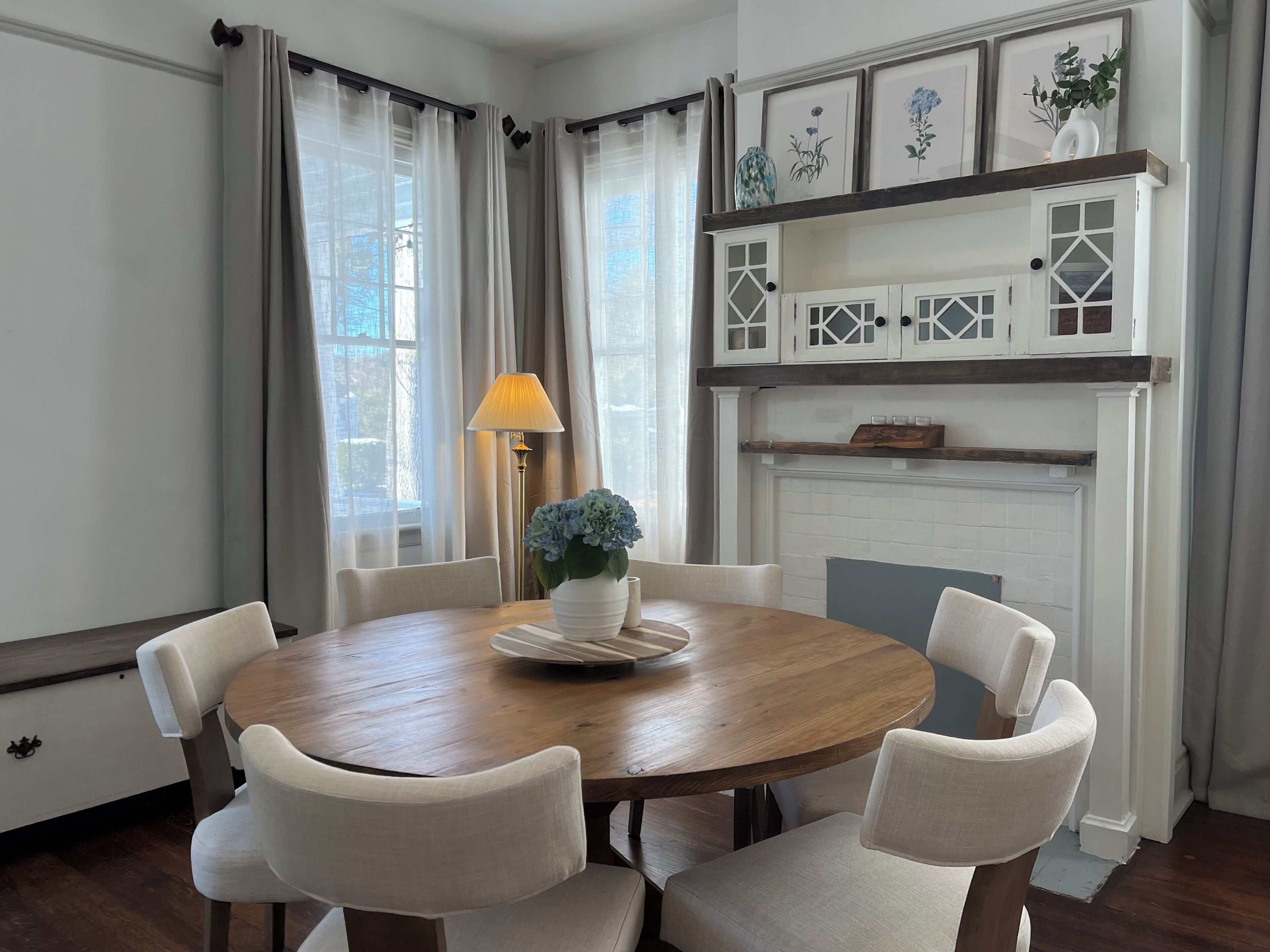 A dining room with a round wooden table, six chairs, a vase with flowers, a lamp, and a fireplace with decorative shelves.