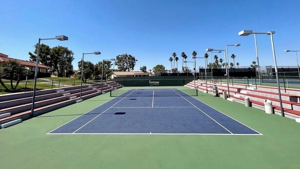 An outdoor tennis court with green and blue surfaces, surrounded by bleachers and street lamps. - Spacious 3BR Desert Condo | Pools, Tennis & Near El Paseo Shopping (Palm Desert)