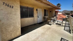 A building with a beige exterior, a number '73062' on the wall, a door, and a patio area with a table and chairs. - Spacious 3BR Desert Condo | Pools, Tennis & Near El Paseo Shopping (Palm Desert)