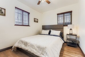 A bedroom with a bed, two windows with blinds, a ceiling fan, and a bedside table with a lamp. - Charming 3-bedroom house in Denver, Quiet with Yard and Easy Commute (Denver)