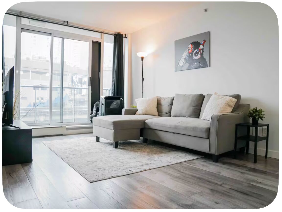 A modern living room with a sectional sofa, a flat-screen TV, a floor lamp, and a large window with a view of buildings.