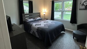 Un dormitorio con cama, escritorio, silla, ventilador de techo, ventana con cortinas y lámpara.