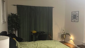 Ein Schlafzimmer mit einem Bett, einem Schreibtisch, einem Stuhl, einer Lampe, einer Pflanze, einem Vorhang, einer Wanduhr und einer Wandleuchte.