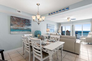 A dining area with a white table and chairs, a chandelier, and a painting of a beach scene. - Waterfront 2BR condo in Panama City Beach, perfect for a relaxing getaway. (Panama City Beach)