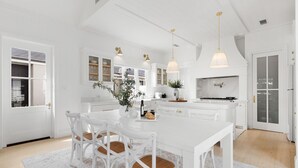 A modern kitchen with white cabinetry, a central island, pendant lighting, and a dining area with white chairs. - The Homestead at Farmhouse Hunter Valley (Pokolbin)