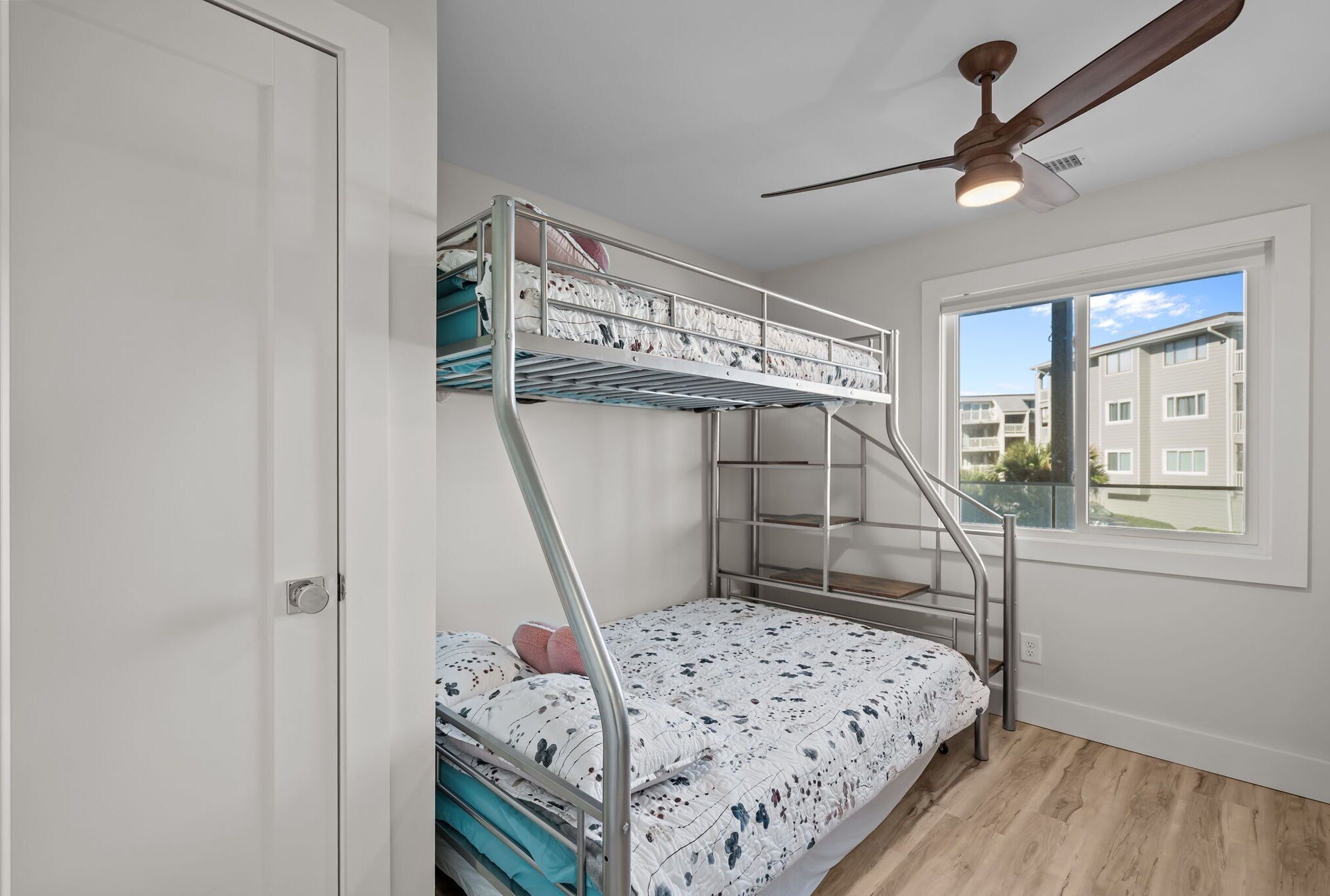 A bunk bed with a metal ladder, a ceiling fan, and a window with a view of buildings.