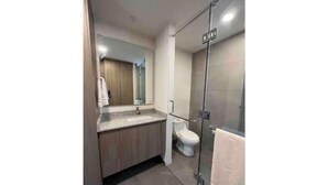 A modern bathroom with a glass shower enclosure, a white toilet, and a wooden vanity with a mirror. - Cozy apartment in Americana 16 (Guadalajara)